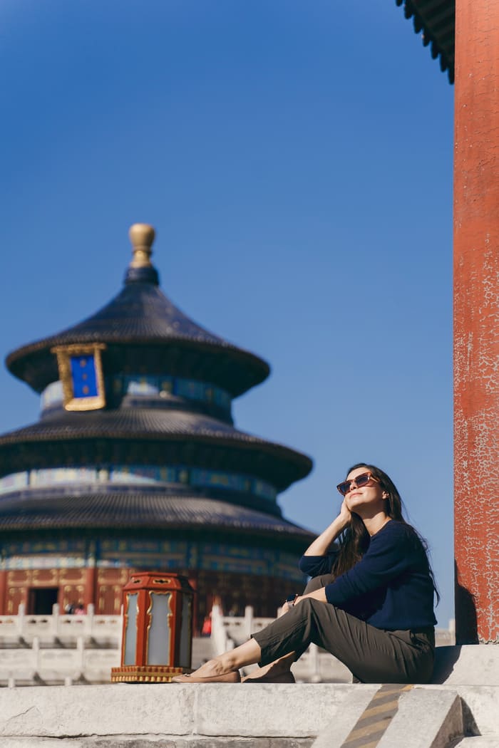 Mujer sentada al sol junto a una columna roja, con el Templo del Cielo de Pekín al fondo bajo un cielo azul despejado.